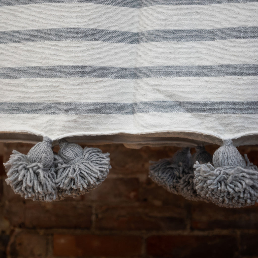 Close up of woven cotton gray and cream striped blanket with gray poms on the edge