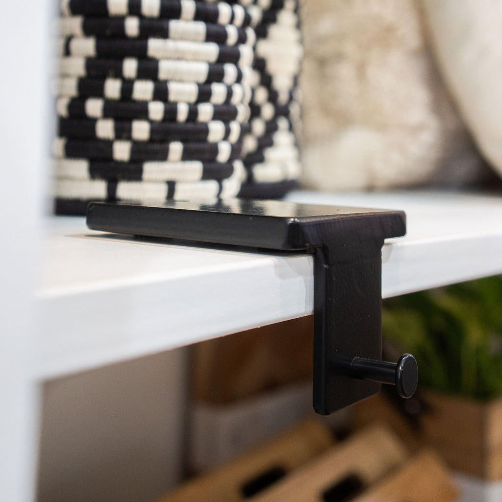 Black iron shelf knob hook resting on large flat weighted back part on white shelf in front of black and white shop background. 