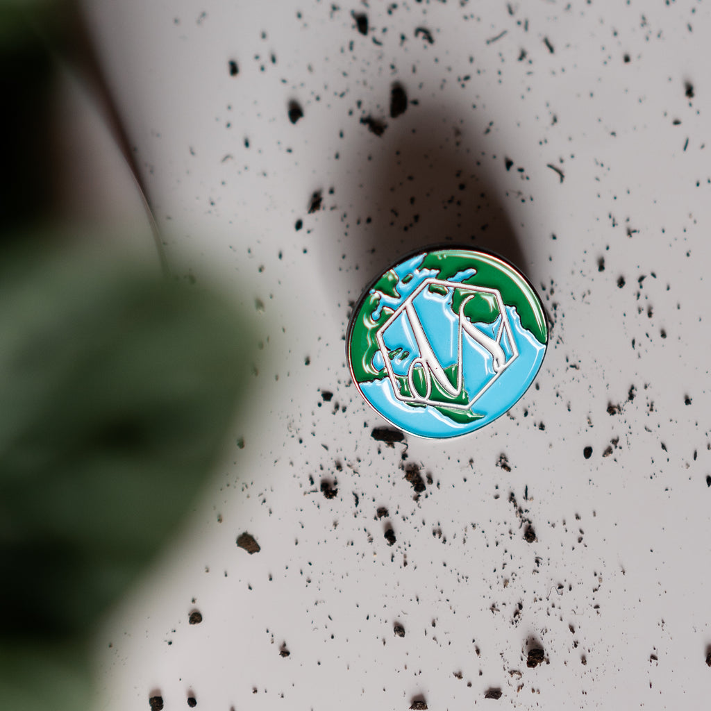 Enamel pin with vintage Driftless Style logo on top of a globe illustration. Pin lays on a dirt filled background.