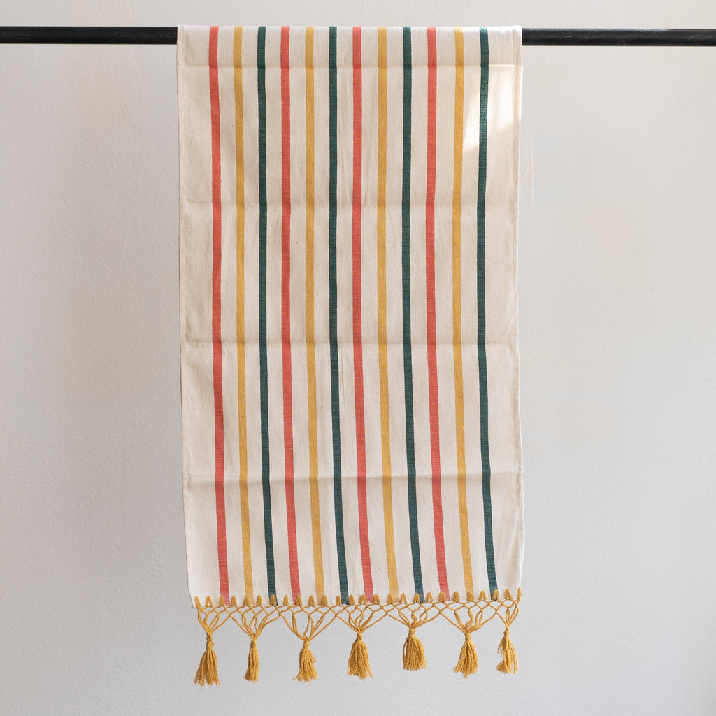 Handwoven cotton table runner in modern Oaxacan striped design. Dark green, mustard, and coral stripes on cream background. Macrame ending in mustard colored knots and tassels on the edge.
