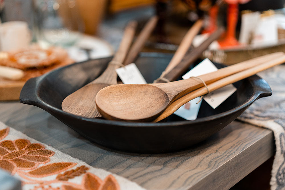Walnut wood spatula and spoon sets rest in a black handled serving bowl on a table.