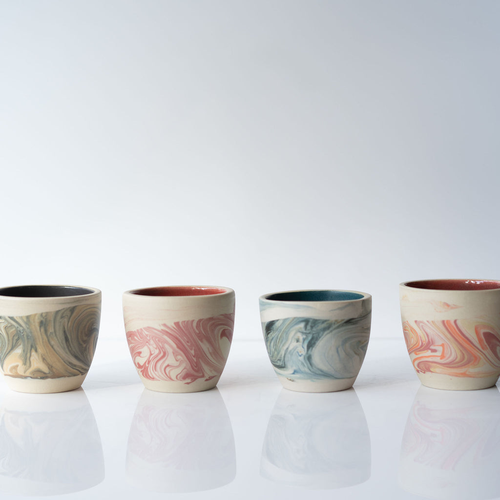 Row of 4 marbled ceramic cups on white background. From left, black and tan marble, pink marble, blues marble, pink and orange marble. The interiors are glazed and finished with a solid color with a raw finish on the exterior. 