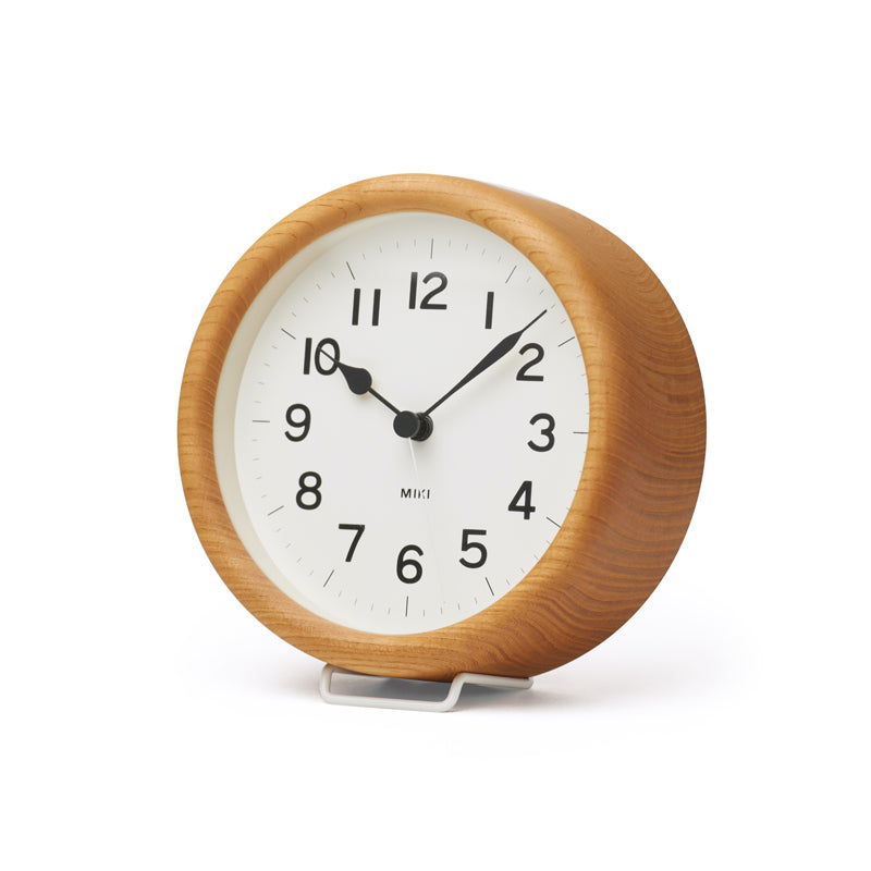 Modern round wood clock with white face and black numbers and hands. Sits on a white background with a little white stand.