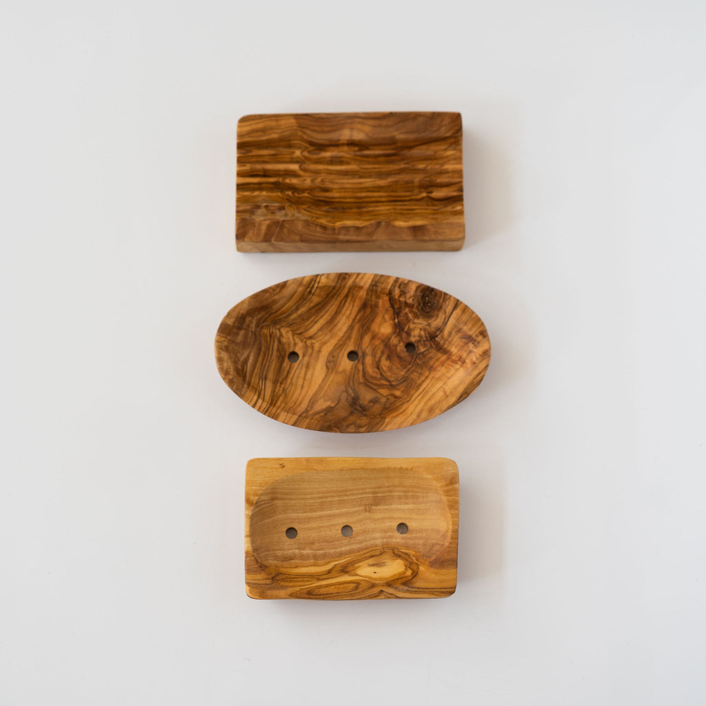 Vertical row of three olive wood soap dishes. Rectangle with draining stripes then oval with drainage holes then rectangle with drainage holes. Light gray background.