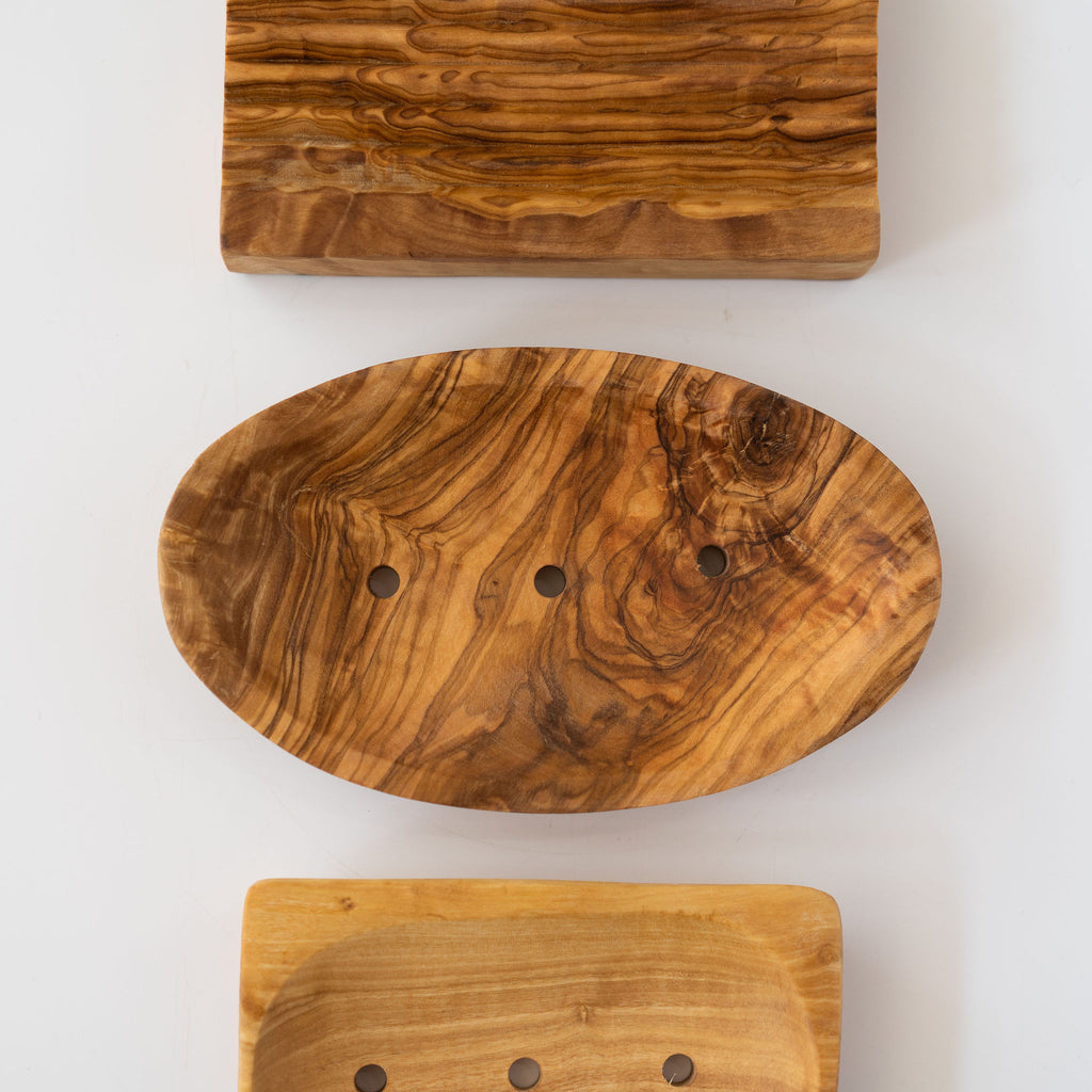 Vertical row of three olive wood soap dishes. Rectangle with draining stripes then oval with a flower soap then rectangle with drainage holes. Light gray background.
