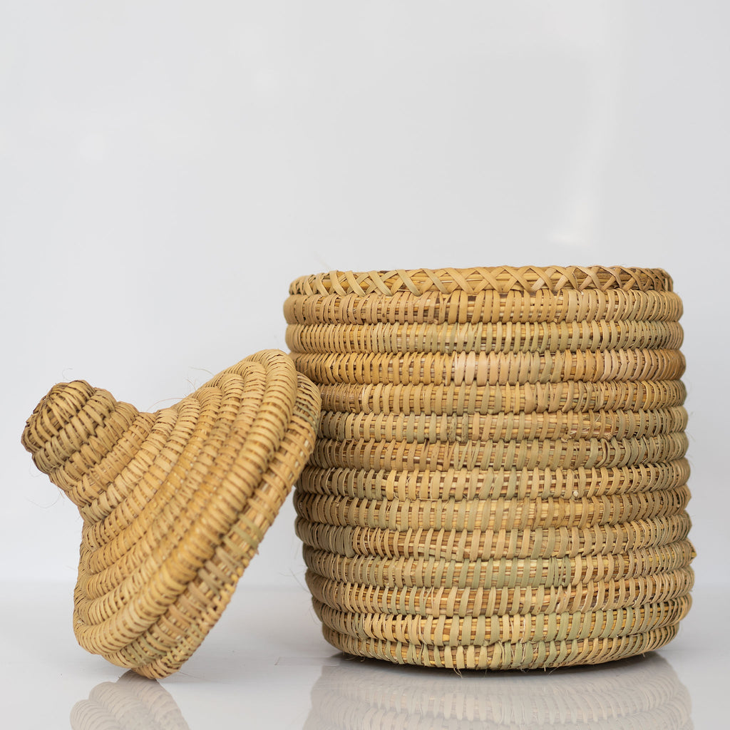 Small rush fiber handwoven basket with lid that has a knob handle. Lid sits next to basket. Light gray background.