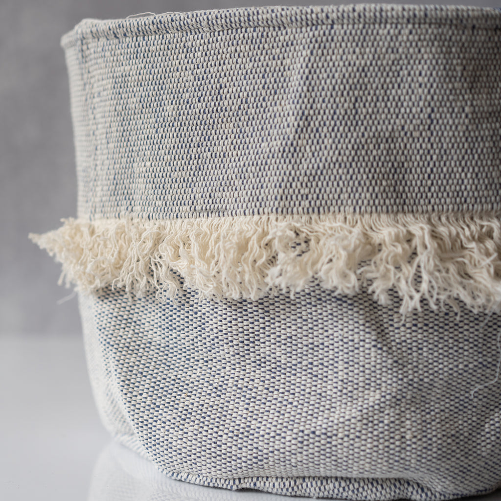 Handwoven cotton denim blue soft sided basket with a strip of white fringe around the middle. Sits on a gray background.
