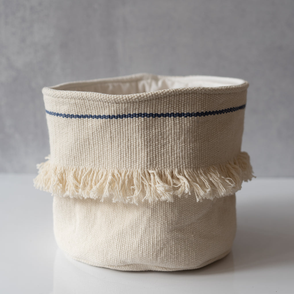 Handwoven cotton cream soft sided basket with an indigo line around the top and a strip of white fringe around the middle. Sits on a gray background.