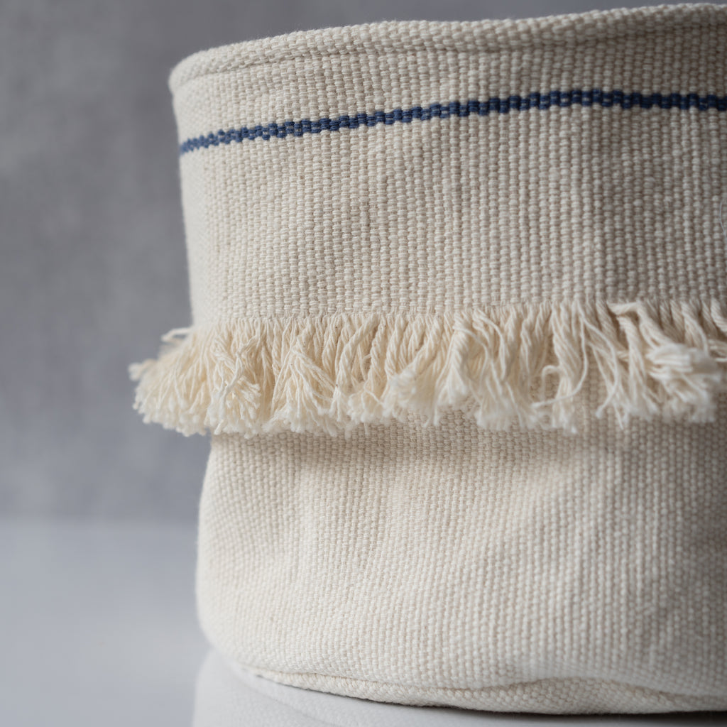 Handwoven cotton cream soft sided basket with an indigo line around the top and a strip of white fringe around the middle. Sits on a gray background.