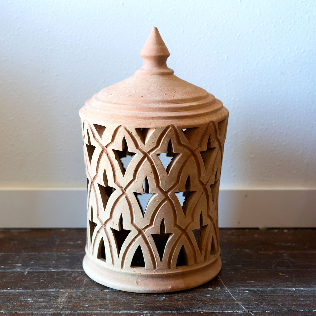 A terra cotta lantern sits on a wood floor in front of a white wall. Traditional punched Moroccan design.