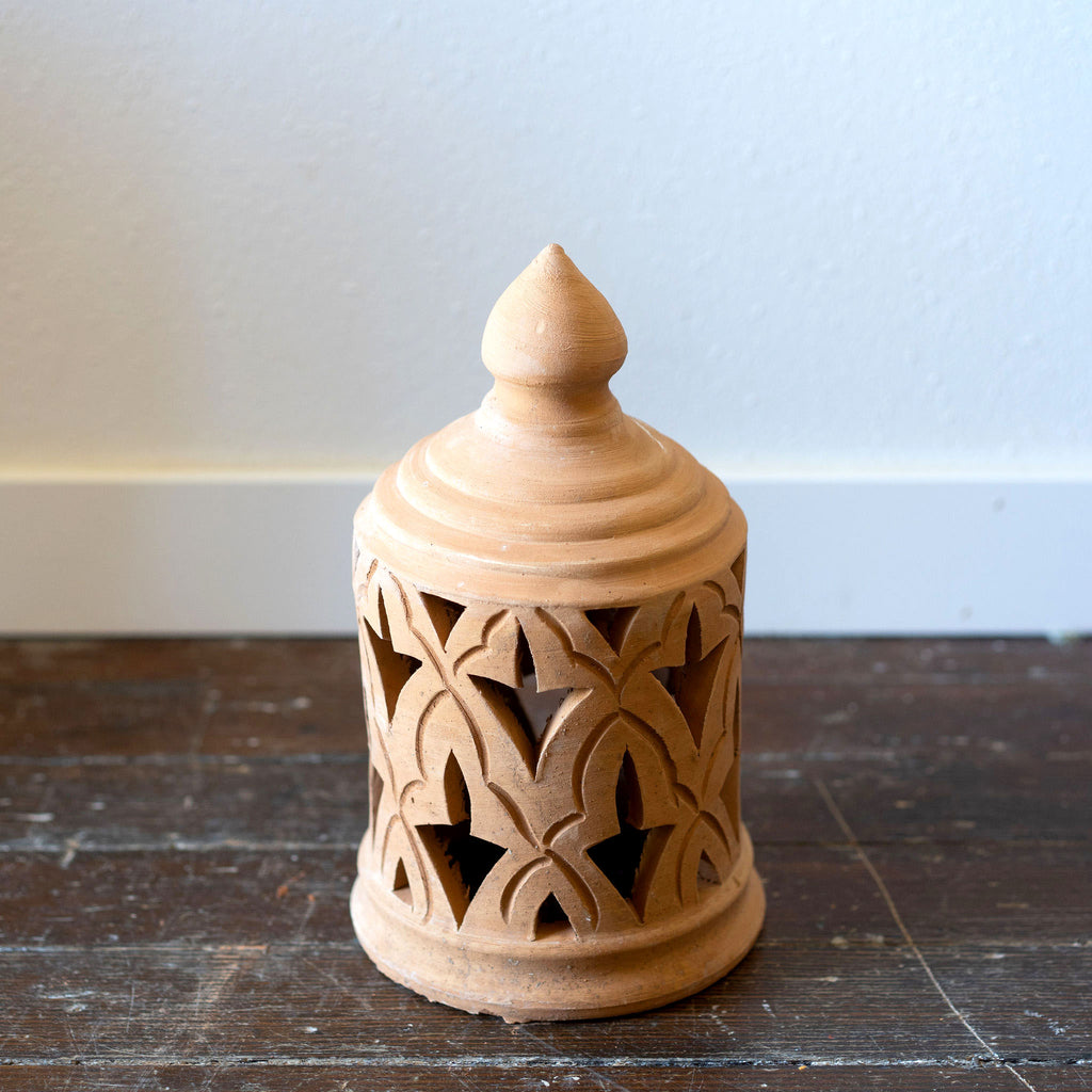 A terra cotta lantern sits on a wood floor in front of a white wall. Traditional punched Moroccan design.