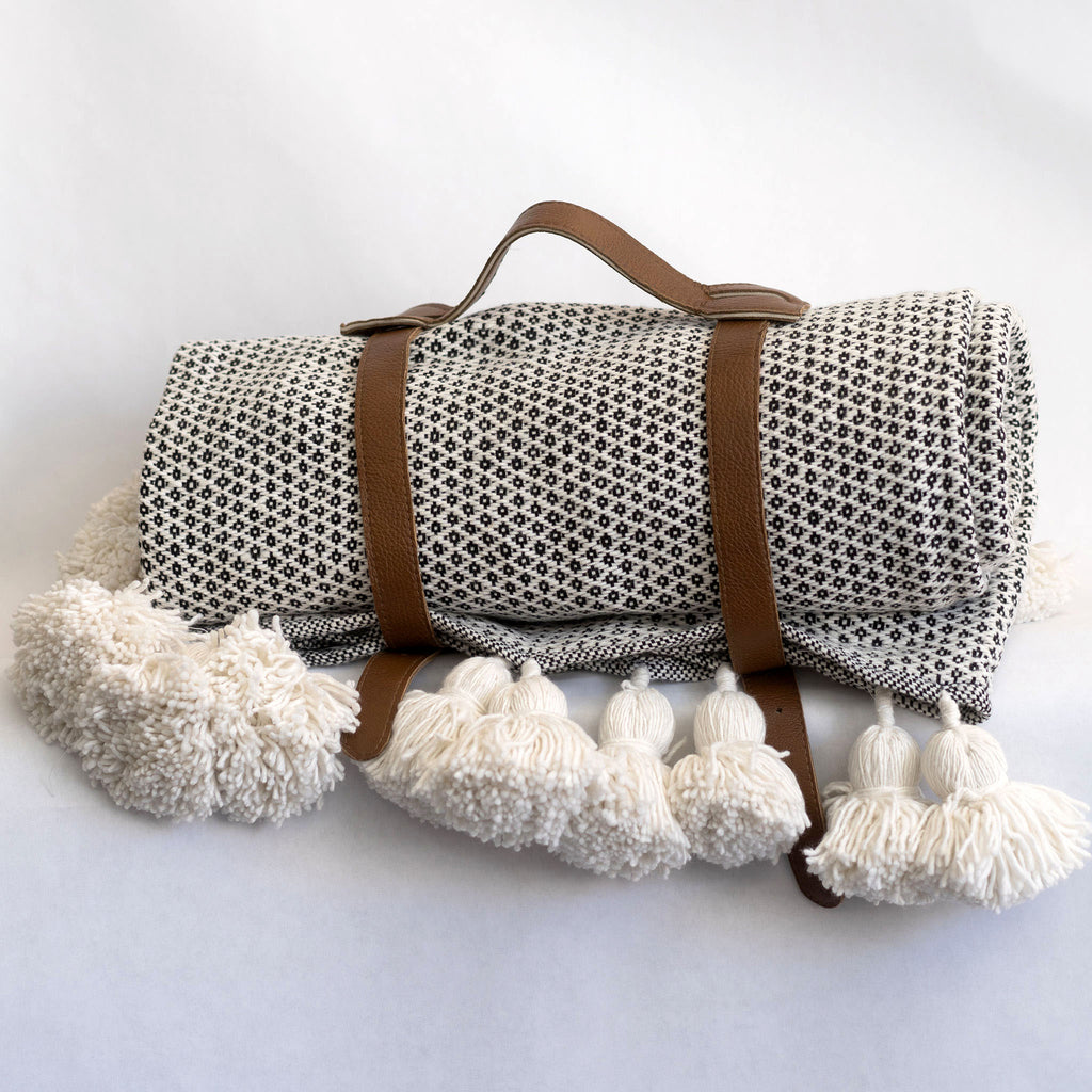 A leather blanket belt with handle wraps around the diamond woven blanket with cream poms. White background.