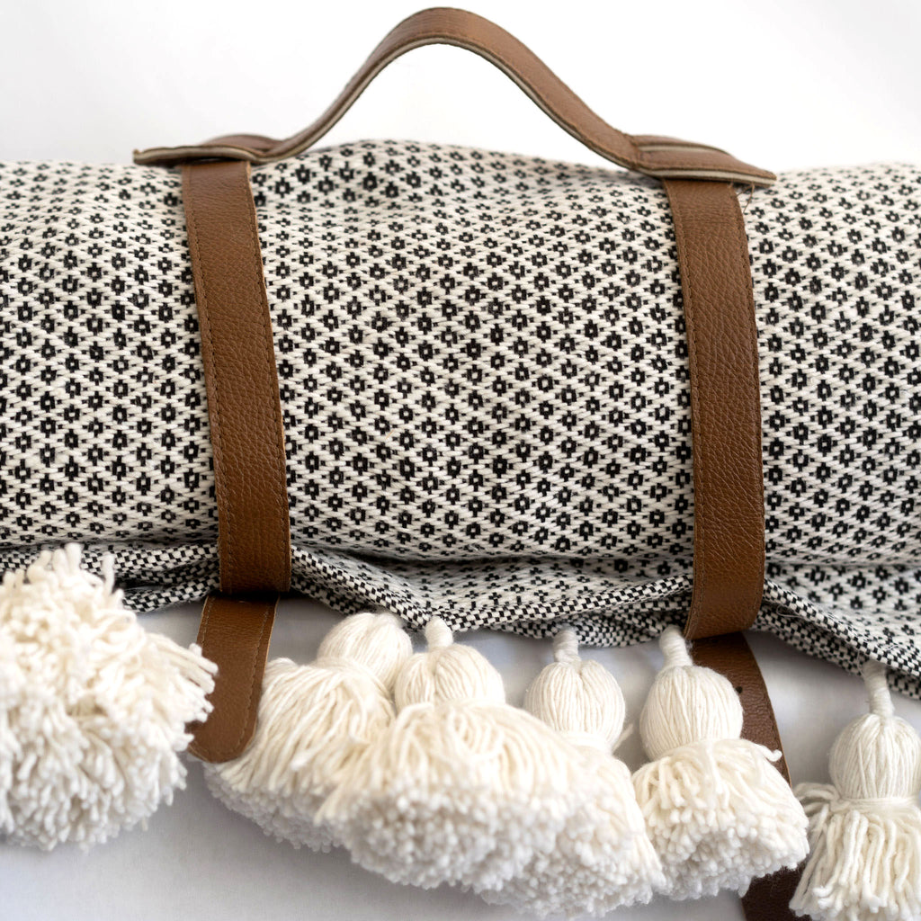 A leather blanket belt with handle wraps around the diamond woven blanket with cream poms.
