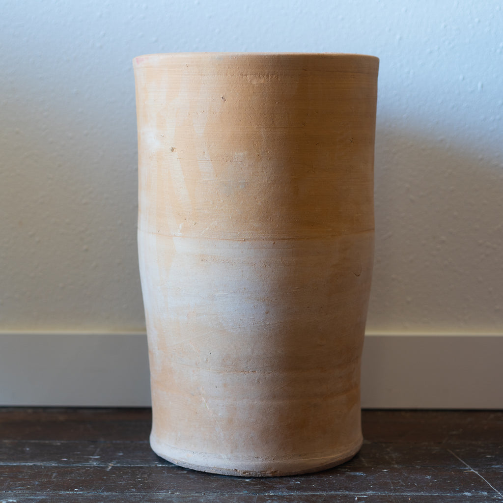Column shaped terra cotta plant pot in front of a white wall sitting on a wood floor.