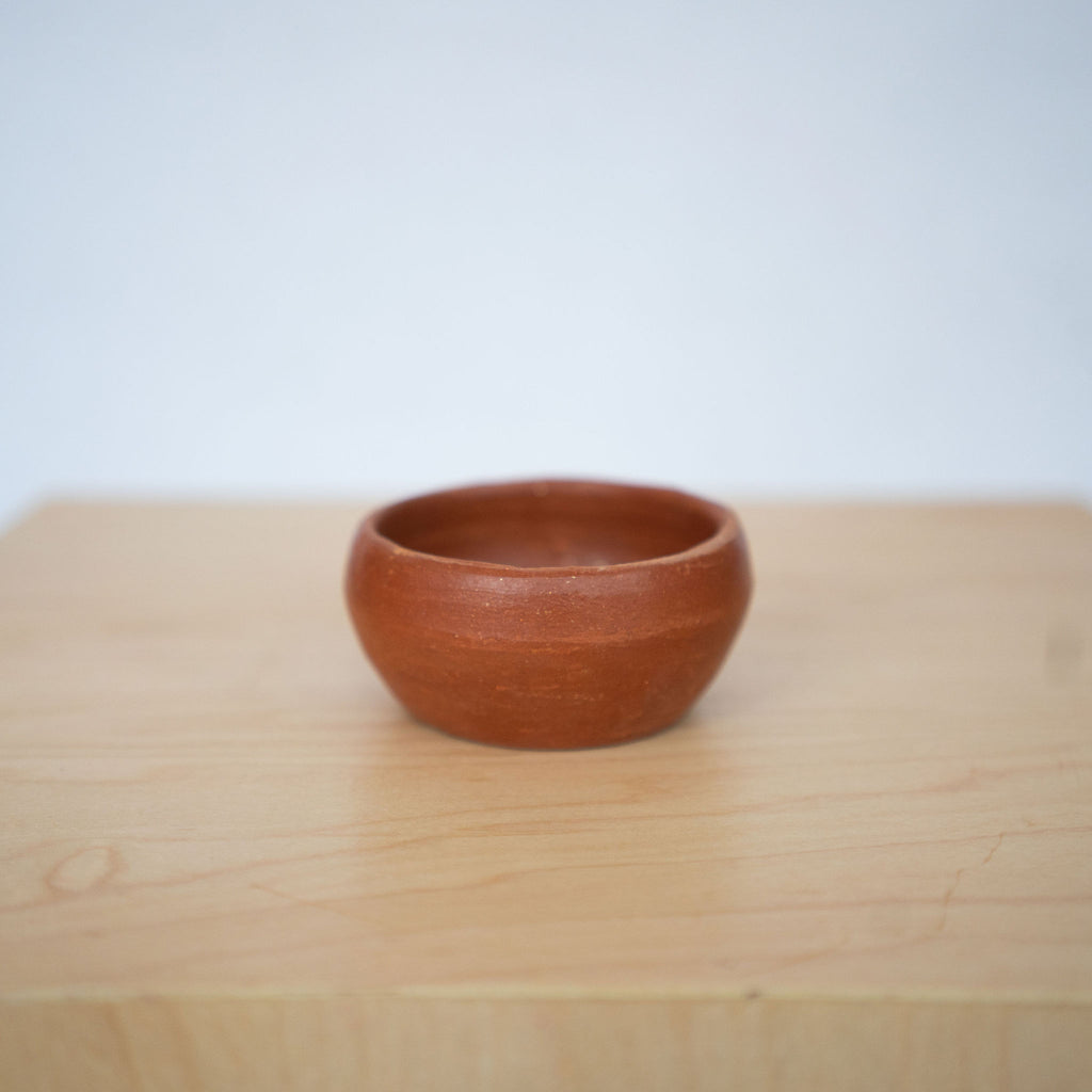 One small rust toned earthenware bowl sits on a wood platform in front of a white background. 