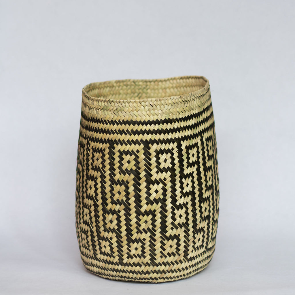 Handwoven palm fiber straight sided basket in traditional Oaxacan designs. Tan and black maze design with squares in diagonal rows with contrasting dots inside each square. Three black stripes at top and bottom edge. Gray background.