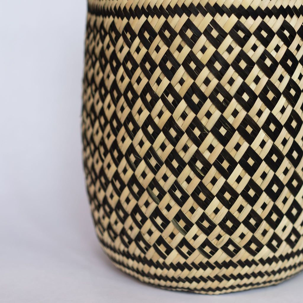 Handwoven palm fiber straight sided basket in traditional Oaxacan designs. Tan and black alternating small diamonds with contrasting dots inside each diamond. Three black stripes at top and bottom edge. Gray background.