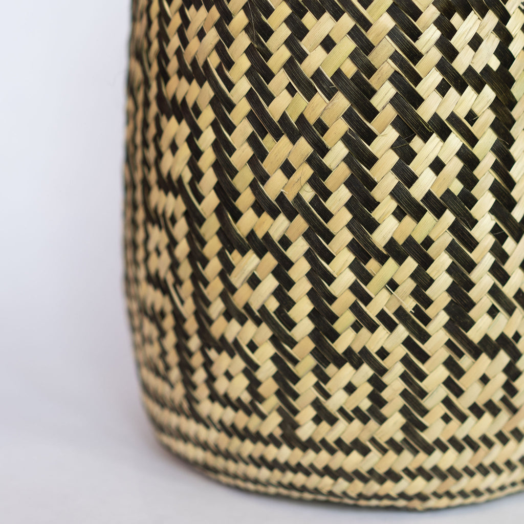 Close up of handwoven palm fiber straight sided basket in traditional Oaxacan designs. Tan and black maze design with squares in diagonal rows with contrasting dots inside each square. Three black stripes at top and bottom edge. Gray background.
