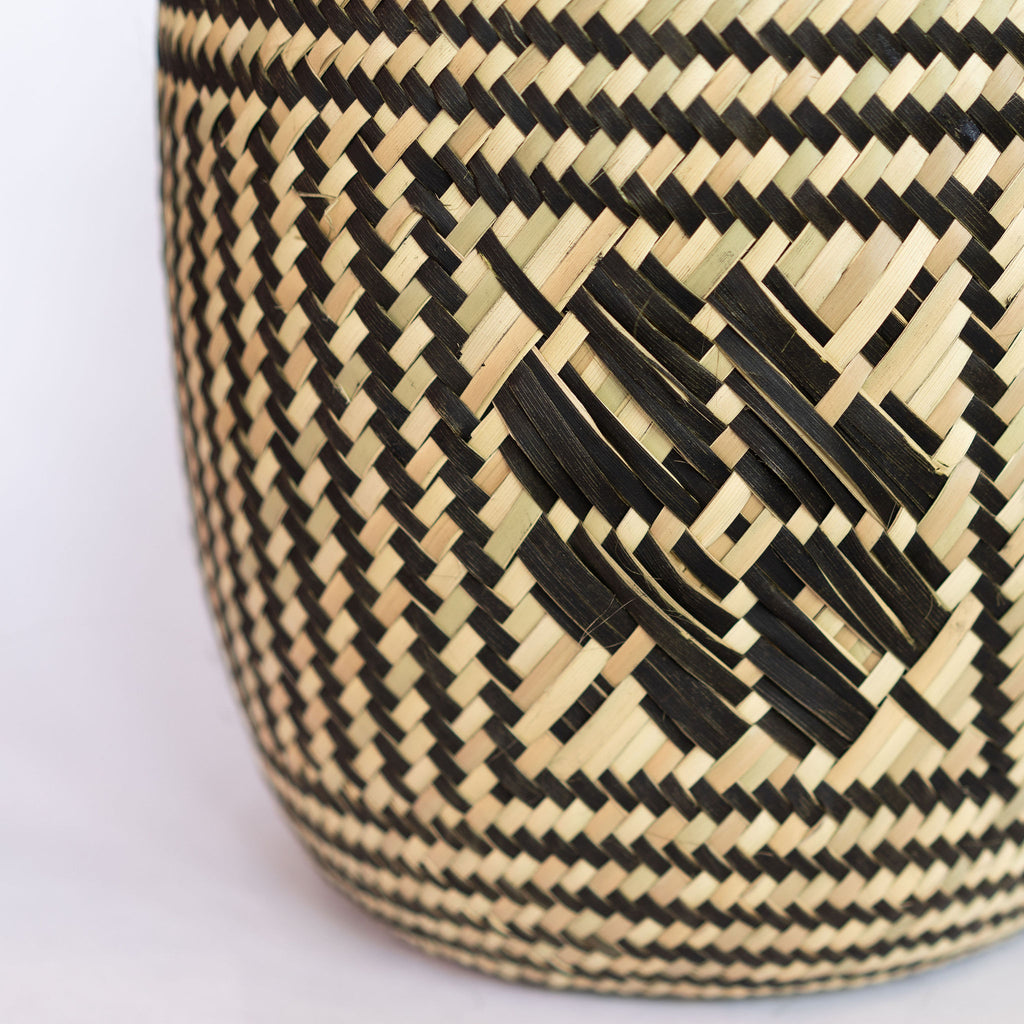 Close up of handwoven palm fiber straight sided basket in traditional Oaxacan designs. Tan and black vertical striped design with flowers on each side. Three black horizontal stripes at top and bottom edge. Gray background.