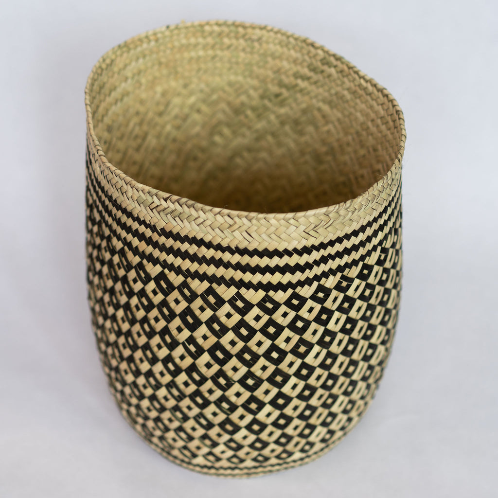 Close up of handwoven palm fiber straight sided basket in traditional Oaxacan designs. Tan and black alternating small diamonds with contrasting dots inside each diamond. Three black stripes at top and bottom edge. Interior is natural tan. Gray background.