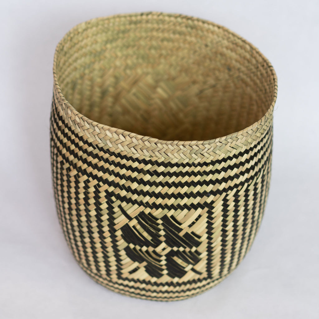 Handwoven palm fiber straight sided basket in traditional Oaxacan designs. Tan and black vertical striped design with flowers on each side. Three black horizontal stripes at top and bottom edge. Interior of basket is natural tan. Gray background.