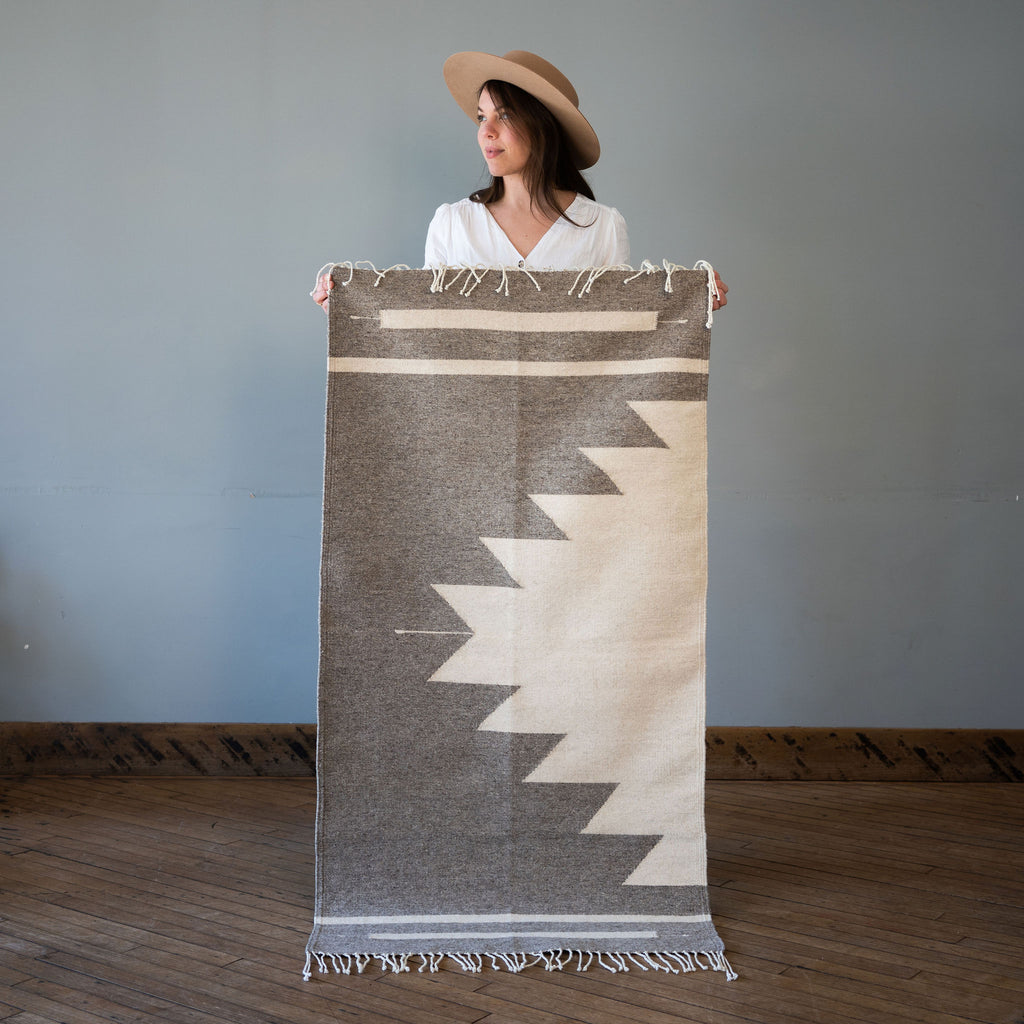 A flat woven wool Oaxacan rug with gray background and cream Aztec-inspired pyramid rising off of one side surrounded by two cream lines on either side. Rug is held up against a grey wall and wood floor by Kelsie in a hat.