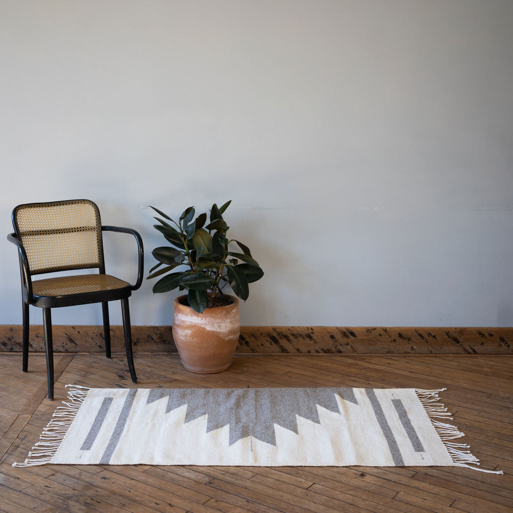 A flat woven wool Oaxacan rug with cream background and gray Aztec-inspired pyramid rising off of one side surrounded by one rattan chair and a potted plant. All in front of a gray wall. Wood floors.