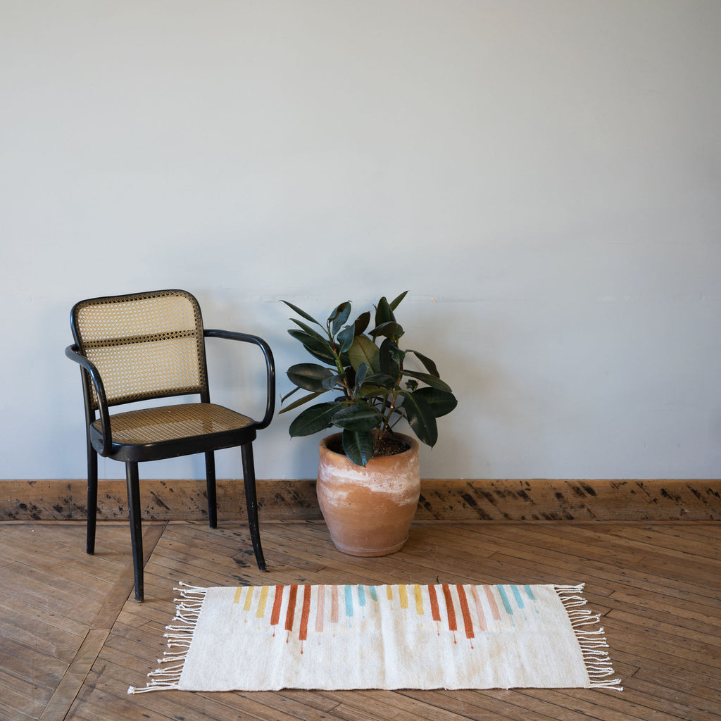 A flat woven cream Oaxacan rug with a geometric triangle made out of lines in Light Blue, Blush, Terracotta, Ochre surrounded by one rattan chair and a potted plant. All in front of a gray wall. Wood floors.
