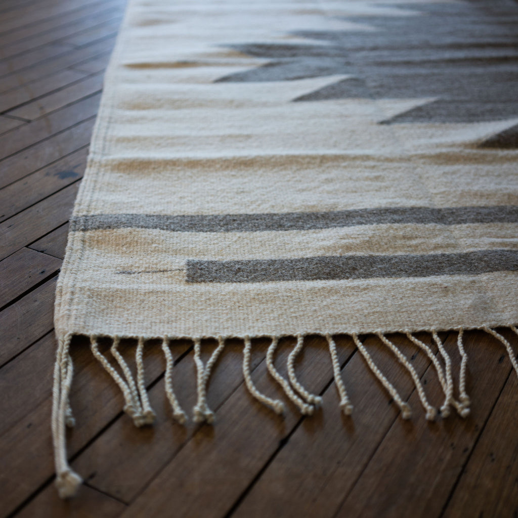 Close up of the fringed corner of a flat woven wool Oaxacan rug with cream back ground and gray Aztec-inspired pyramid rising off of one side. Wood floor.