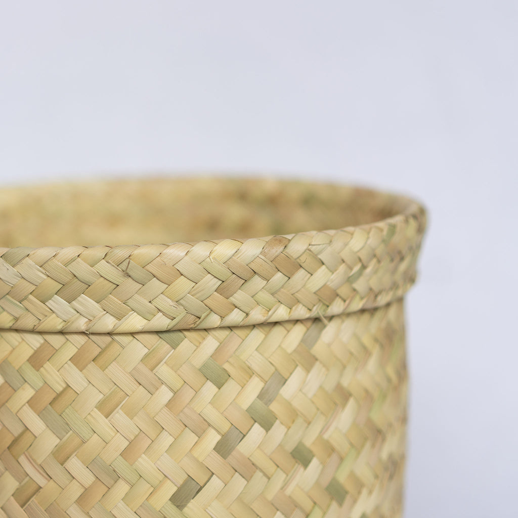 Close up of a short handwoven palm fiber straight sided basket in natural tan. Slight ridge around top. Gray background. 
