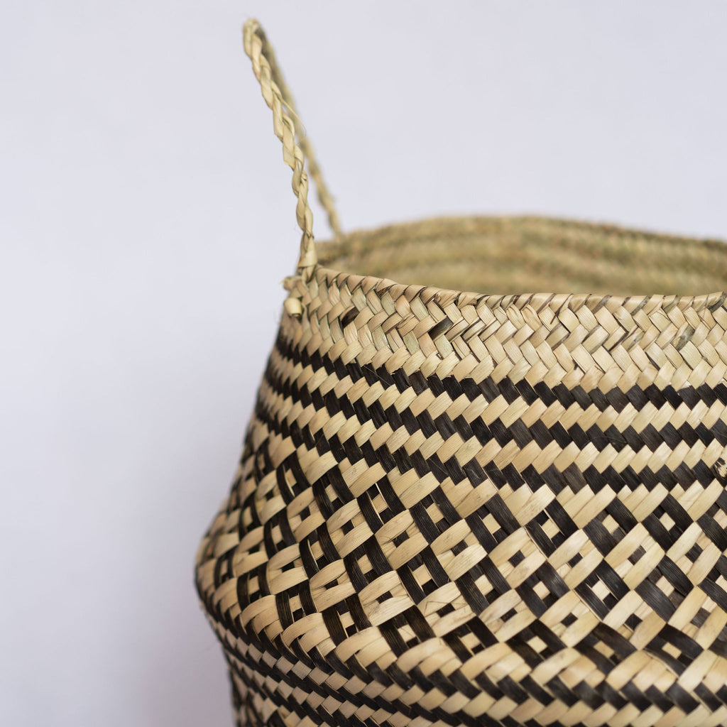 Close up of handle of handwoven palm fiber belly basket in traditional Oaxacan designs. Tan and black alternating small diamonds with contrasting dots inside each diamond. Three black stripes at the middle and at the top and bottom edge. Gray background.