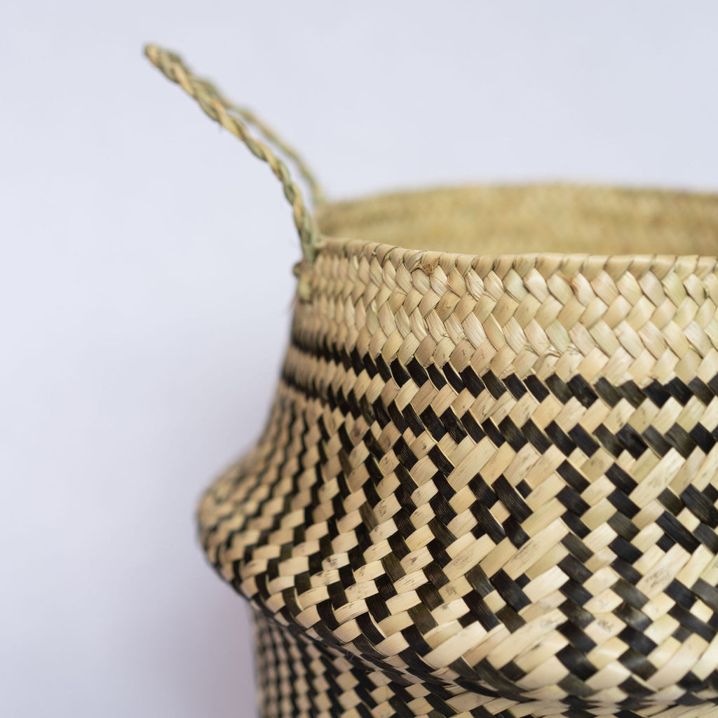 Close up of handle of handwoven palm fiber belly basket in traditional Oaxacan designs. Tan and black vertical stripes alternate with two vertical rows of small squares in each quarter of the basket.  Horizontal stripes run along both top and bottom edge. Gray background.