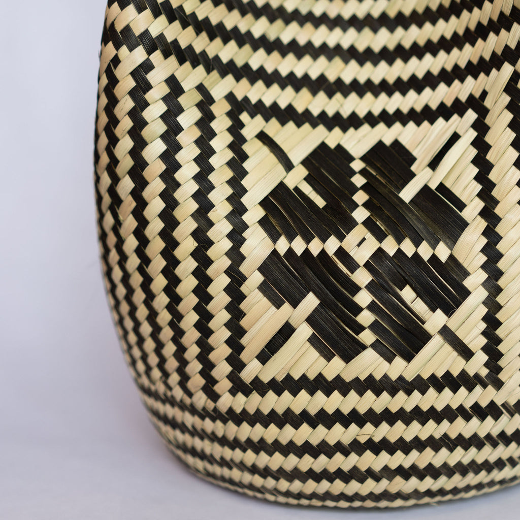 Close up of a handwoven palm fiber tote bag with black leather handles in traditional Oaxacan designs. Tan and black vertical striped design with flowers on each side. Three black horizontal stripes at top and bottom edge. Gray background.