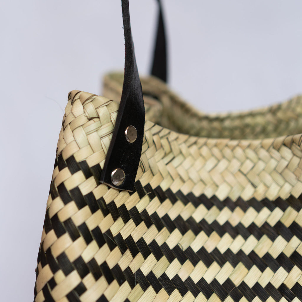 Close up of the handle attachment to a handwoven palm fiber tote bag with black leather handles in traditional Oaxacan designs. Tan and black vertical striped design with flowers on each side. Three black horizontal stripes at top and bottom edge. Gray background.