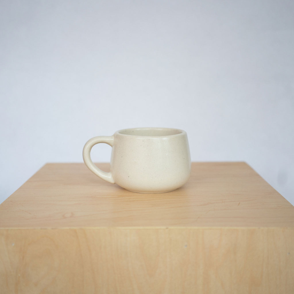 White round short ceramic mug sits on a wood platform in front of a light gray background.