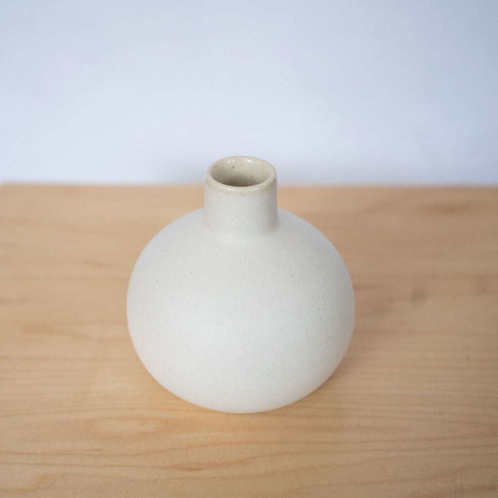 A small round white ceramic vase with skinny neck sits on top of a wood platform in front of a light gray background.