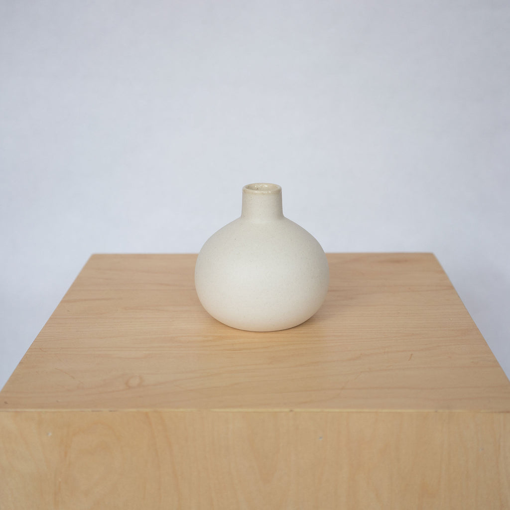 A small round white ceramic vase with skinny neck sits on top of a wood platform in front of a light gray background.
