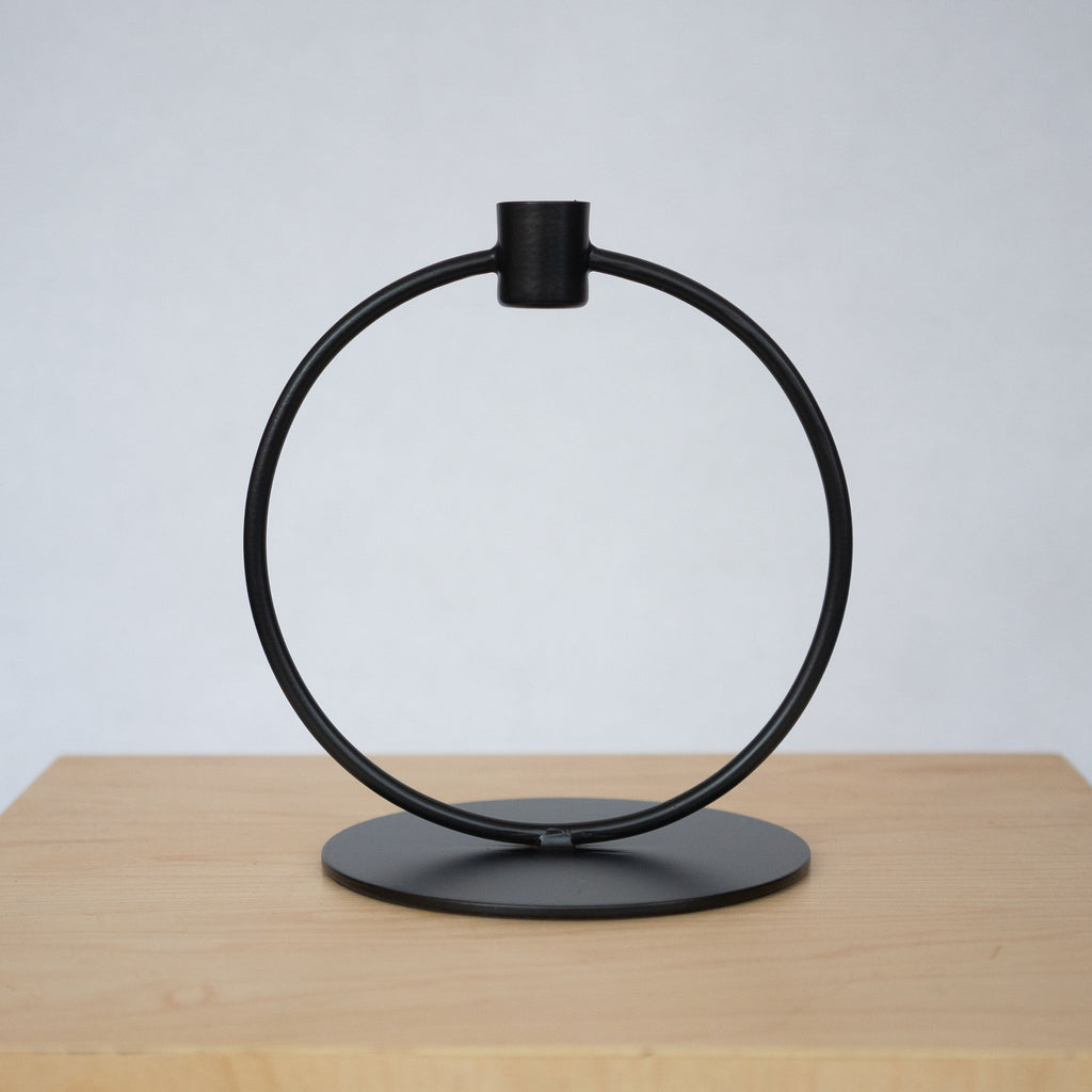 Powder coated iron black large circle taper candle holder with a flat round base and a small cup for the candle at the top. Sits on a wood platform in front of a light gray background.