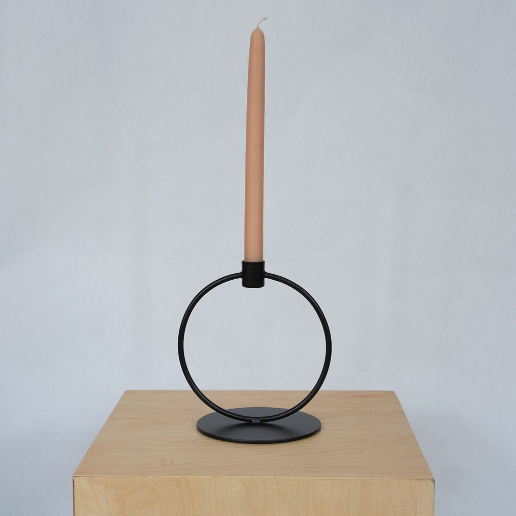 Powder coated iron black large circle taper candle holder with a flat round base and a small cup with a peach taper candle at the top. Sits on a wood platform in front of a light gray background.
