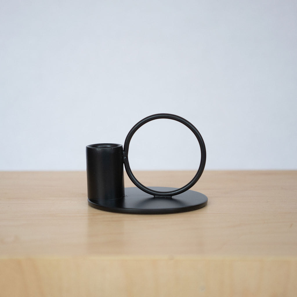 Powder coated iron black small taper candle holder with a circle set next to a cup for a taper candle all on a small round base. Both sit on a wood platform in front of a light gray background.