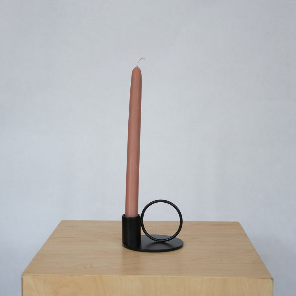 Powder coated iron black small taper candle holder with a circle set next to a cup with a terra cotta toned taper candle all on a small round base. Both sit on a wood platform in front of a light gray background.