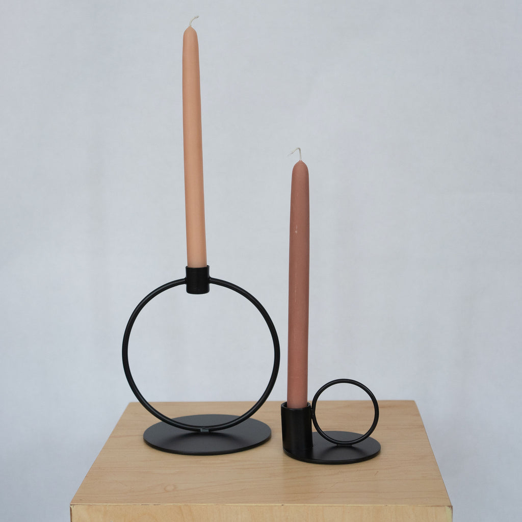 Powder coated iron black taper candle holders. On the left is a large circle with a flat round base and a small cup with a peach taper candle at the top. On the right is a small circle set next to a cup with a terra cotta toned taper candle all on a small round base. Both sit on a wood platform in front of a light gray background.
