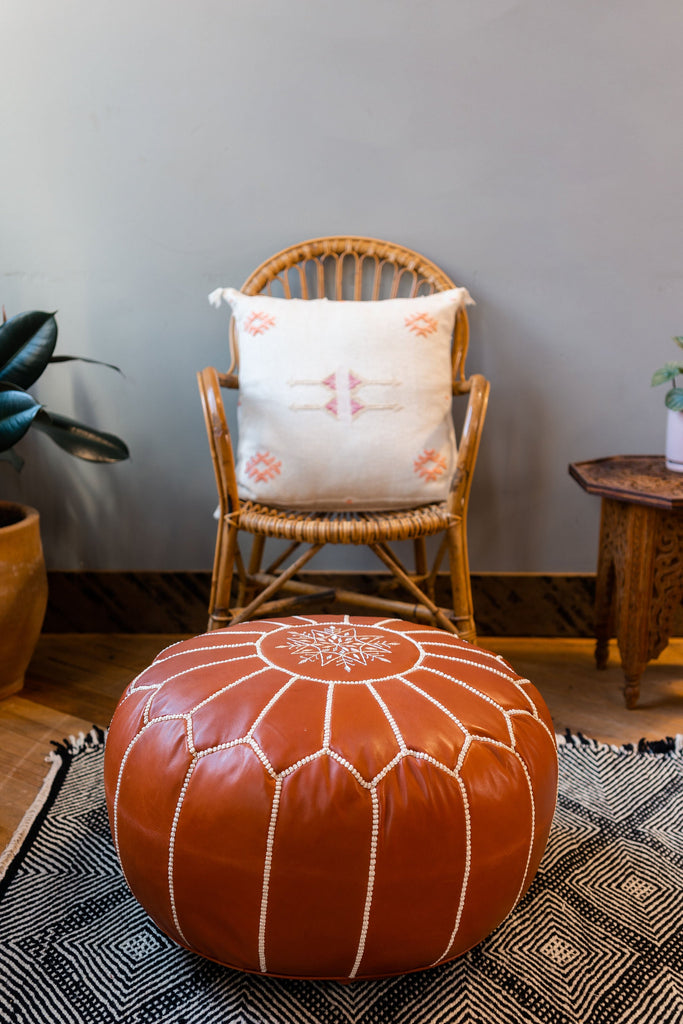 Round Camel Colored Leather Pouf / Ottoman sitting on a Zanafi black and white geometric rug in front of a rattan chair with an ivory pillow. A plant peaks in on one side of the photo. A carved Moroccan side table peaks in on the other side of the photo.