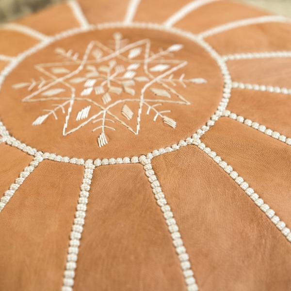 Close up of the round natural leather pouf / ottoman. The embroidery design in the middle of the top is symmetrical, radial design, reminiscent of a snowflake.
