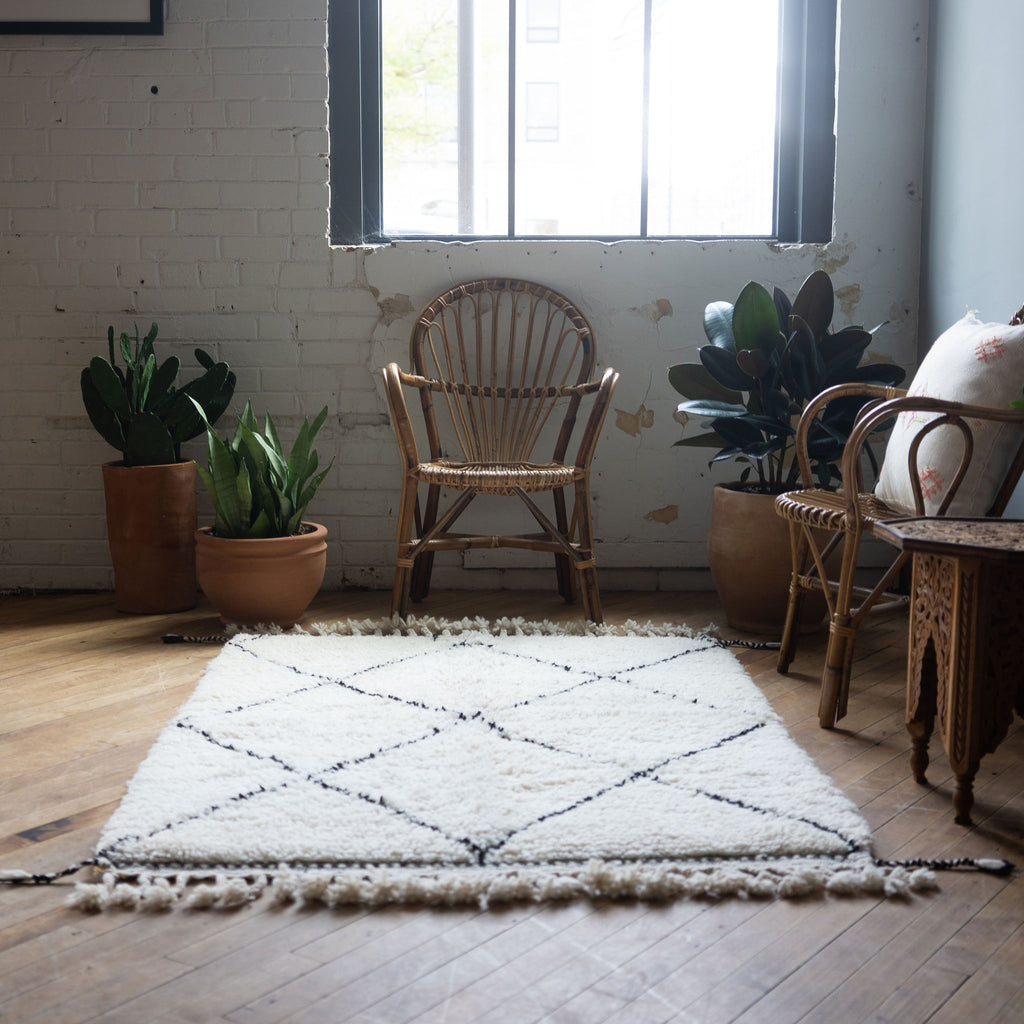 A straight-on view of a high pile Moroccan Beni Ourain rug with classic cream and black diamond design and tasseled/fringed edge surrounded by two rattan chairs, a side table, and a potted plant. Wood floors.