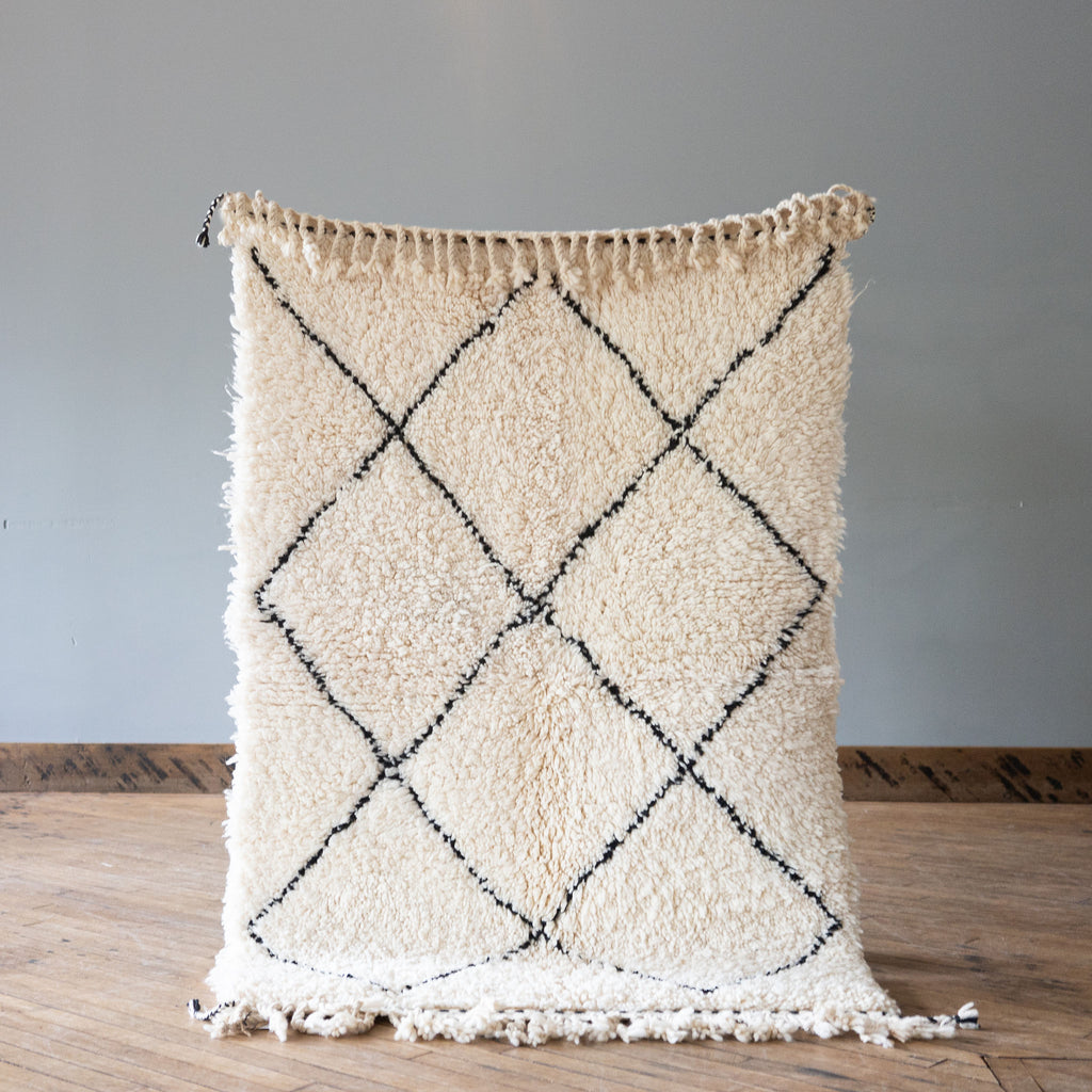 A high pile Moroccan Beni Ourain rug with classic cream and black diamond design and tasseled edge. Rug is held up against a grey wall and wood floor.
