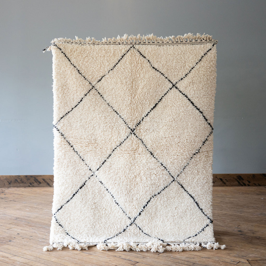 A high pile Moroccan Beni Ourain rug with classic cream and black diamond design and tasseled/ fringe edges. Rug is held up against a grey wall and wood floor.