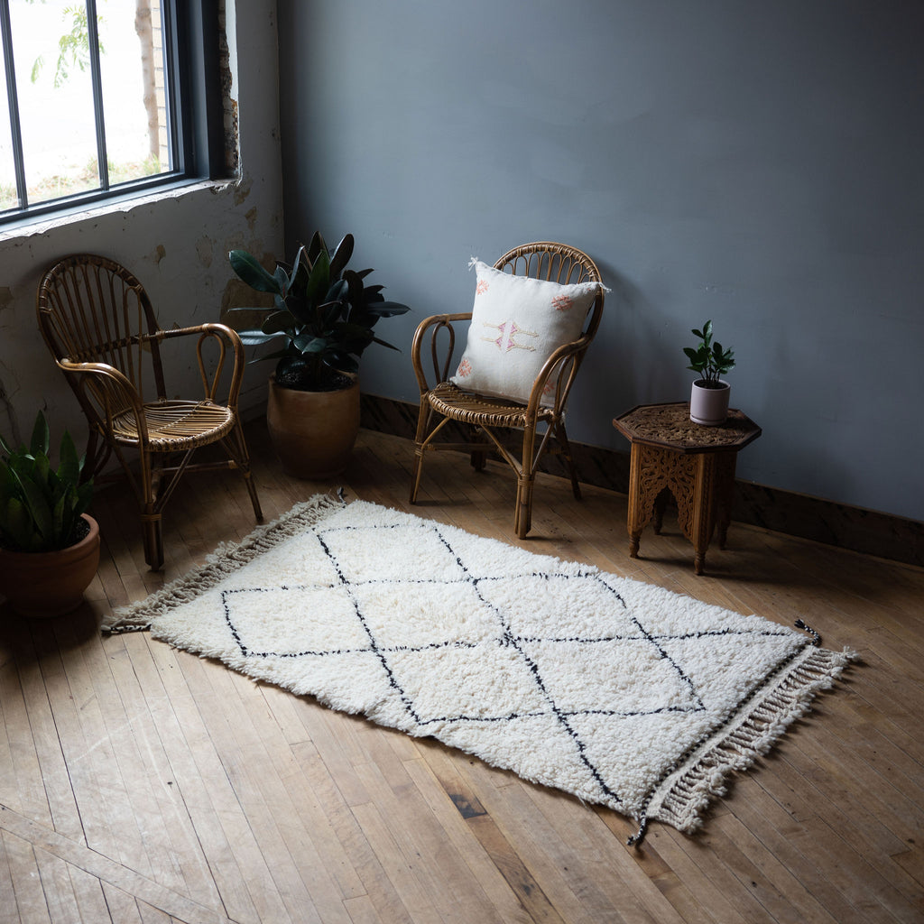 A diagonal view of a high pile Moroccan Beni Ourain rug with classic cream and black diamond design and tasseled/fringed edge surrounded by two rattan chairs, a side table, and a potted plant. Wood floors.