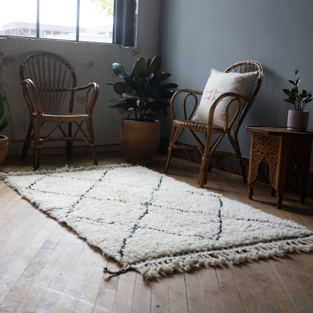 A diagonal view of a high pile Moroccan Beni Ourain rug with classic cream and black diamond design and tasseled/fringed edge surrounded by two rattan chairs, a side table, and a potted plant. Wood floors.