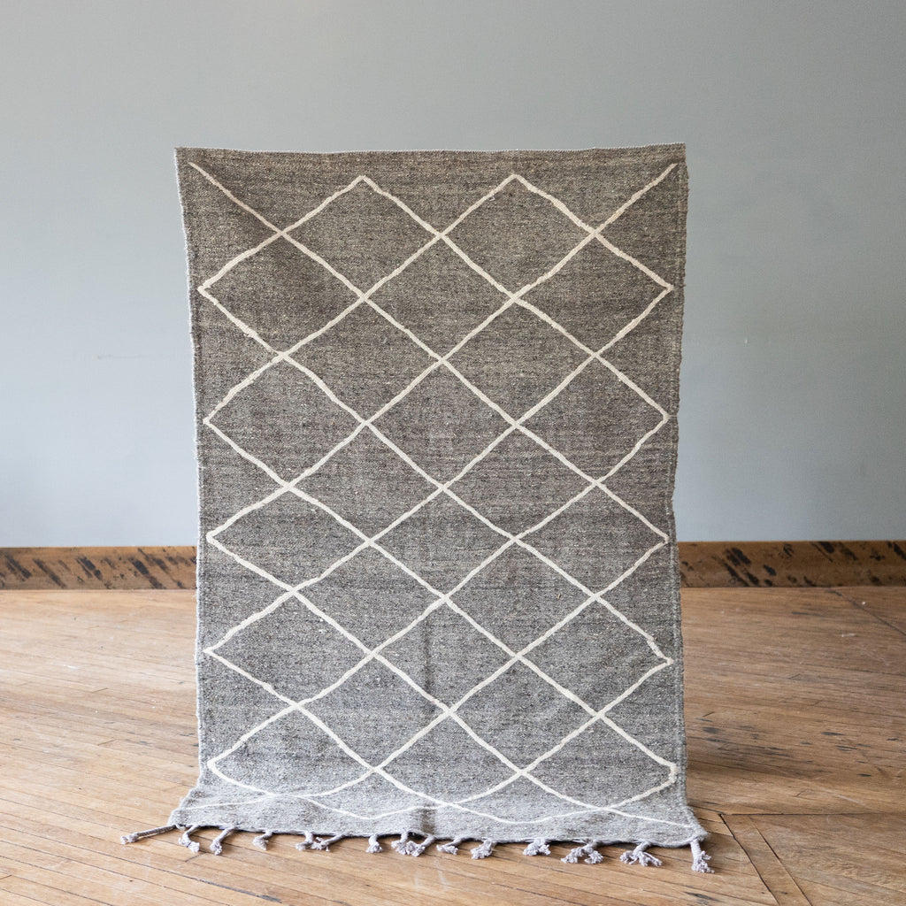 A flat woven wool Moroccan Beni Ourain rug with gray background and cream diamond design. Rug is held up against a grey wall and wood floor.